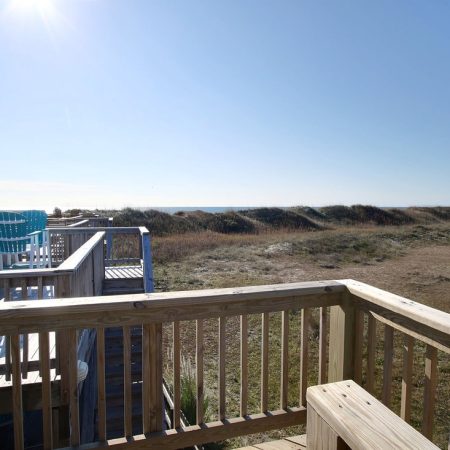 Wooden deck overlooking grassy sand dunes under a clear blue sky with a few chairs visible on an upper level.