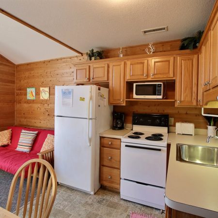 Small wooden kitchen with white appliances, a red futon, dining table, and upper and lower cabinets.