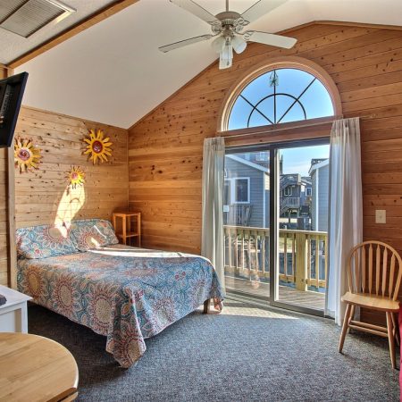 Wood-paneled bedroom with a bed, TV, chairs, and glass doors leading to a balcony with a neighborhood view.