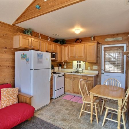 Small wooden kitchen and dining area with a fridge, stove, table, chairs, and a red futon.