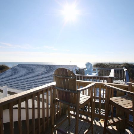 Wooden deck with chairs overlooking rooftops and ocean under a bright sunny sky.