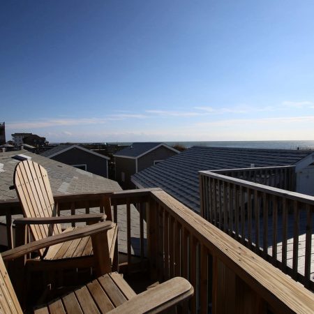 Two wooden chairs sit on a deck overlooking rooftops and the ocean under a clear, sunny sky.