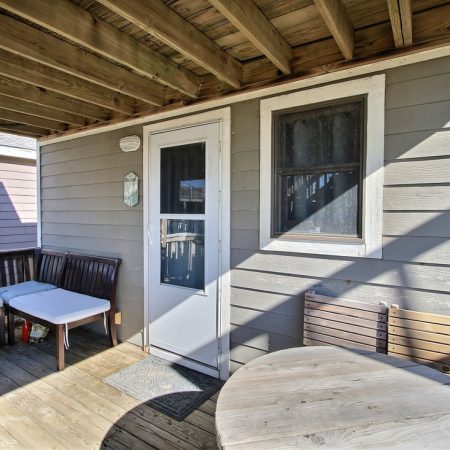 Covered wooden deck with a cushioned bench, round table, wooden chairs, and a door leading inside the house.