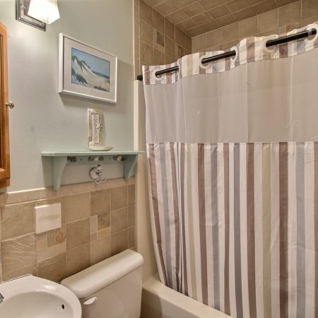 Small bathroom with tiled walls, a toilet, sink, striped shower curtain, wall shelf, and framed picture above the toilet.