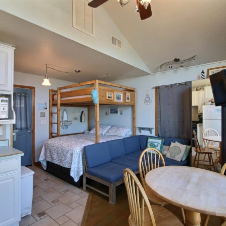 Small studio apartment with a bunk bed, blue sofa, round dining table, wall-mounted TV, and compact kitchen area.