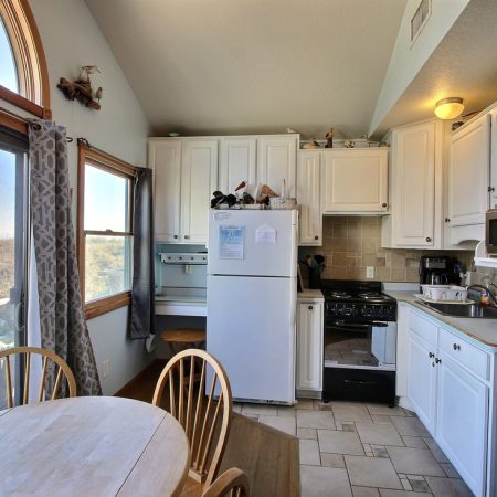 Compact kitchen with white cabinets, a round dining table, wooden chairs, and large windows letting in natural light.