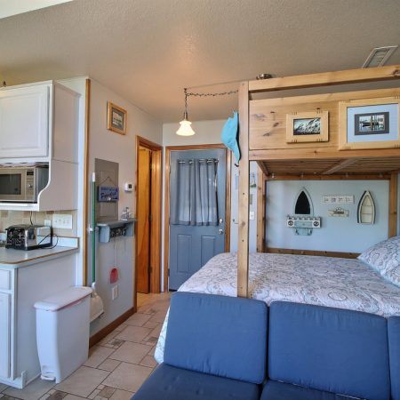 Small studio with a kitchen, bunk bed, blue sofa, wall art, and framed photos, featuring tile flooring.
