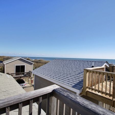 View of rooftops, wooden deck with chairs, and ocean in the distance under a clear blue sky.