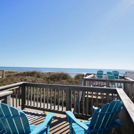 Wooden deck with blue Adirondack chairs overlooks grassy dunes and ocean under a clear, sunny sky.