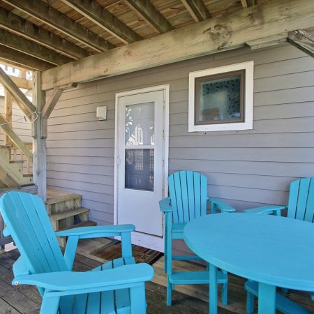 Covered wooden deck with four turquoise chairs around a table, next to a white door and stairs.