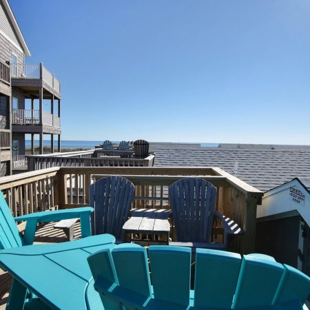 A rooftop deck with teal and gray Adirondack chairs overlooks rooftops and the ocean under a clear blue sky.