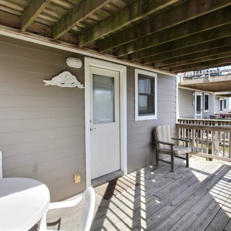 Wooden porch with a white door, small table and chairs, and a cloud-shaped wall decoration under a deck.