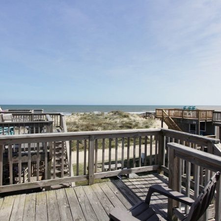Wooden deck with chairs overlooks sand dunes, a path, and the ocean under a clear sky.