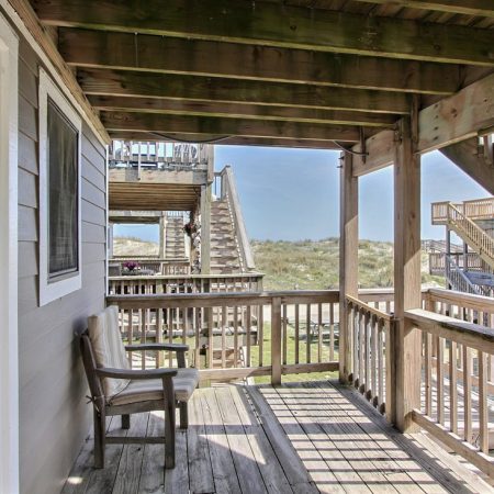 Wooden deck with a bench, stairs, and railings, overlooking grassy dunes and nearby beach houses under a clear sky.