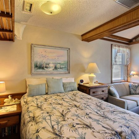 Bedroom with a double bed, nightstands, lamps, a sofa, wall art, and wooden accents, lit by natural and artificial light.