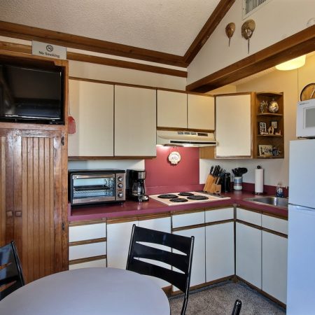 Small kitchen with white cabinets, red countertops, appliances, and a round table with black chairs in the foreground.