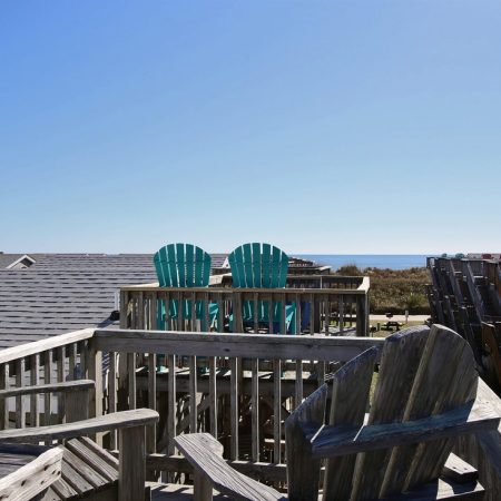 Wooden deck with several chairs overlooks rooftops and the distant ocean under a clear blue sky.