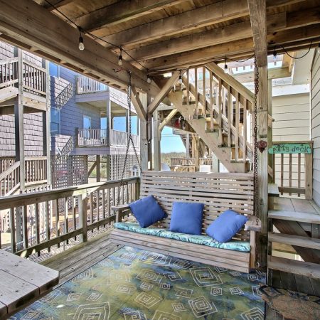 Covered wooden porch with a swing bench, three blue pillows, patterned rug, stairs, and nearby multi-story decks.