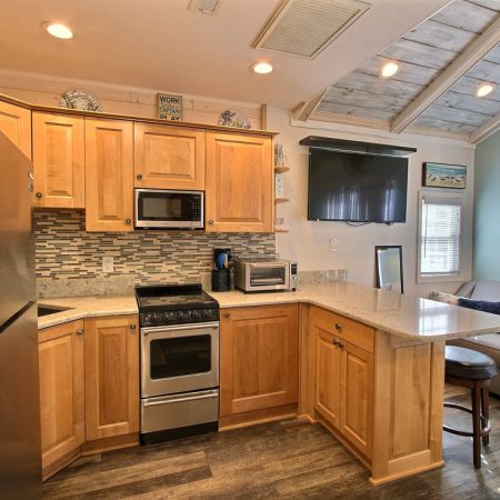 Modern kitchen with wood cabinets, stainless steel appliances, and a small living area with a sofa and TV in the background.