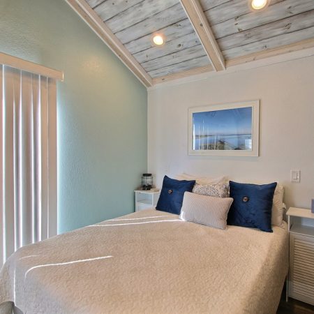 Small bedroom with a white bed, blue accent pillows, side tables, wall art, and vertical blinds letting in sunlight.