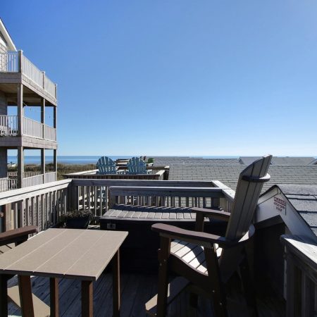 Deck with Adirondack chairs and table overlooks rooftops and the ocean under a clear, sunny blue sky.