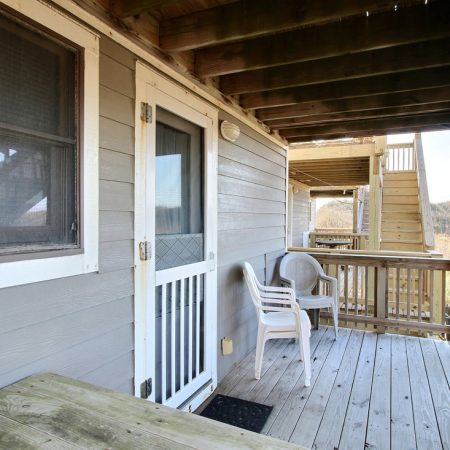 Wooden porch with two white chairs, a table, and railing overlooking grassy scenery under a covered roof.