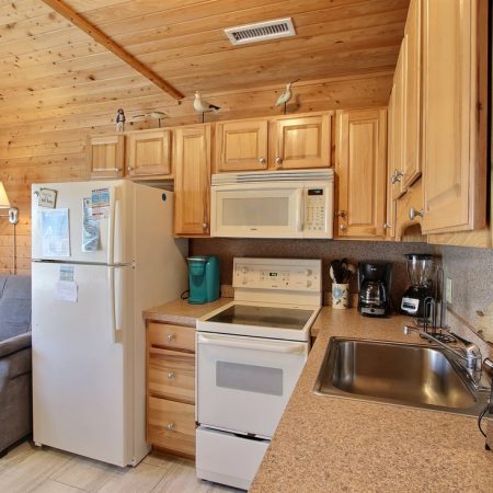 Kitchen with wood cabinets, white stove/fridge, microwave, coffee makers, toaster, sink, sofa, and wood-paneled walls.