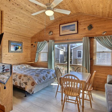 Small wooden cabin interior with a bed, dining table, couch, ceiling fan, windows, and simple ocean-themed decor.
