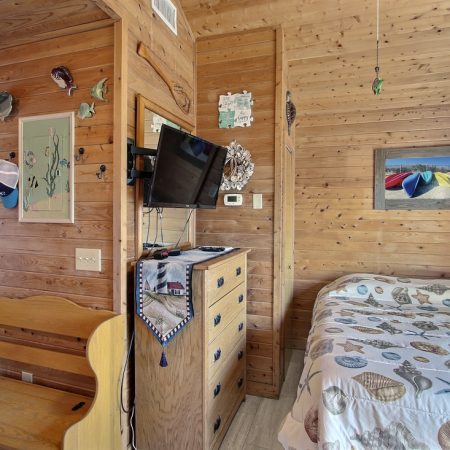 Wood-paneled bedroom with a bed, dresser, wall-mounted TV, wooden bench, and nautical-themed decor.