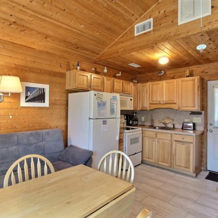 Small wooden cabin kitchen with light wood cabinets, white appliances, a dining table, gray sofa, and wall decor.