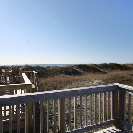 Wooden deck with chairs overlooks grassy sand dunes under a clear blue sky near the ocean.