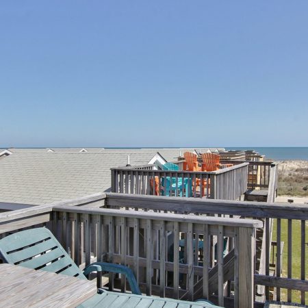 Wooden deck with colorful chairs overlooks rooftops, grassy area, sandy dunes, and the ocean under a clear sky.