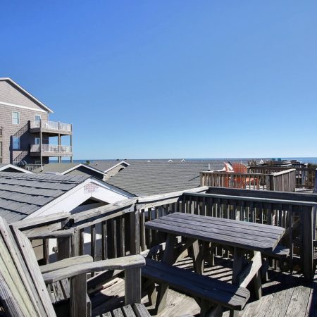 Wooden deck with picnic tables and chairs overlooks rooftops, dunes, and ocean under a clear blue sky.